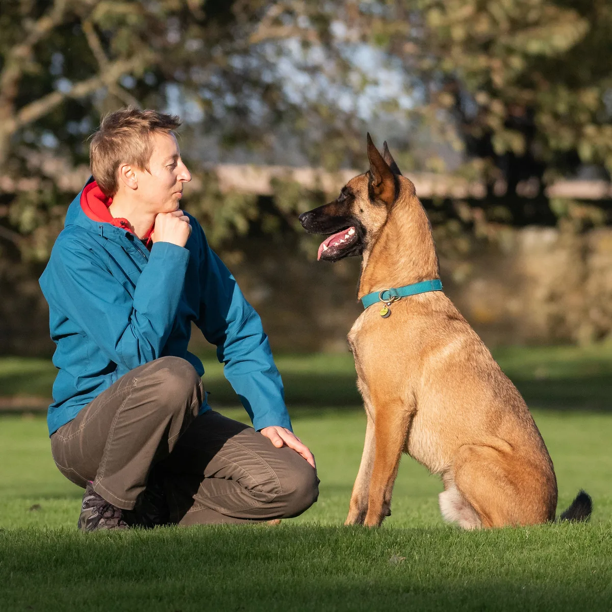 Agata kneeling on the grass beside her Malinois, Arco, who sits looking up at her