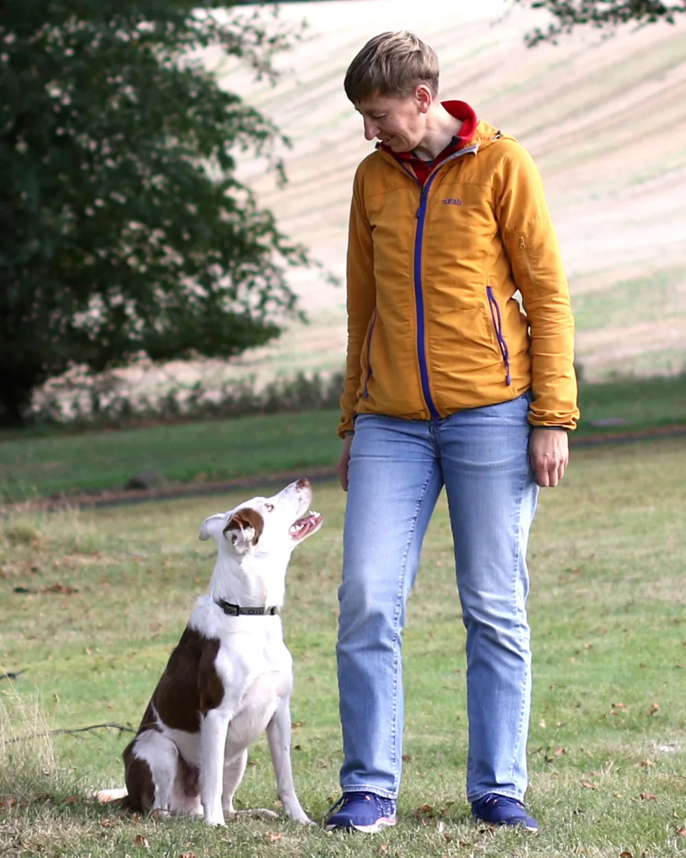 Agata standing in a field with her dog Merrick, who sits beside her looking up at her